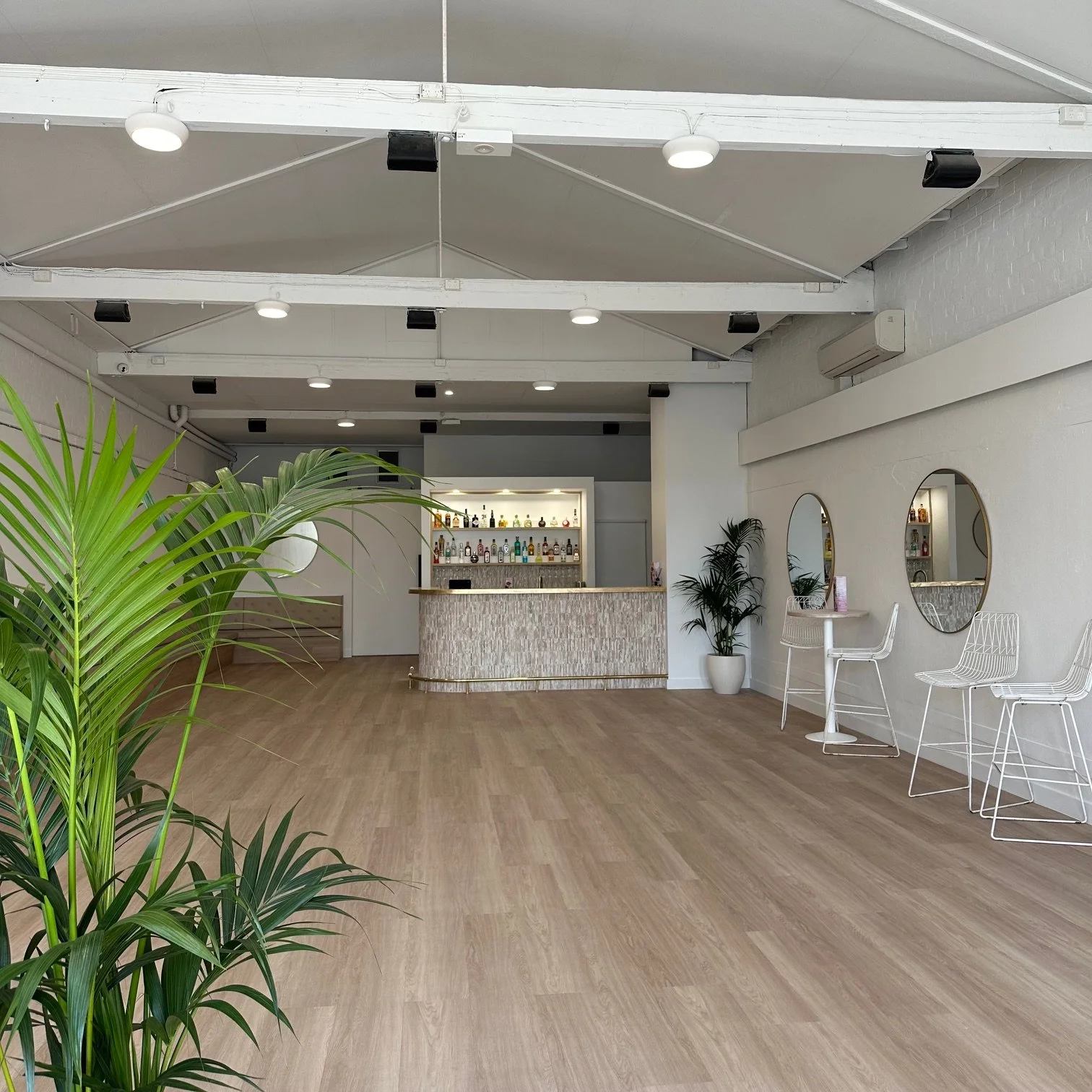 A modern, minimalistic bar with a light wood floor, white walls, and green plants. There is a curved bar counter with bottles on shelves behind it, and seating with white wire chairs and mirrors on the wall.