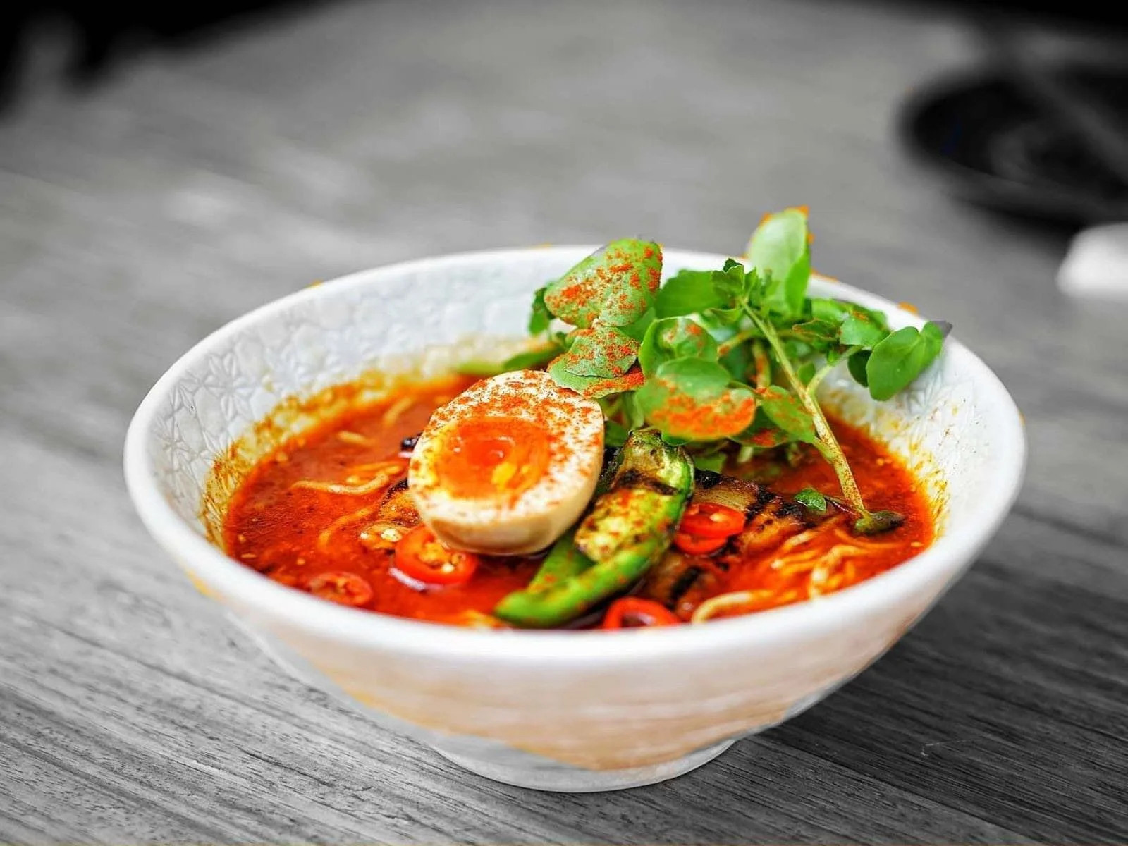 Bowl of spicy Asian noodle soup garnished with fresh herbs, half boiled egg, chili peppers, and vegetables on a wooden table.