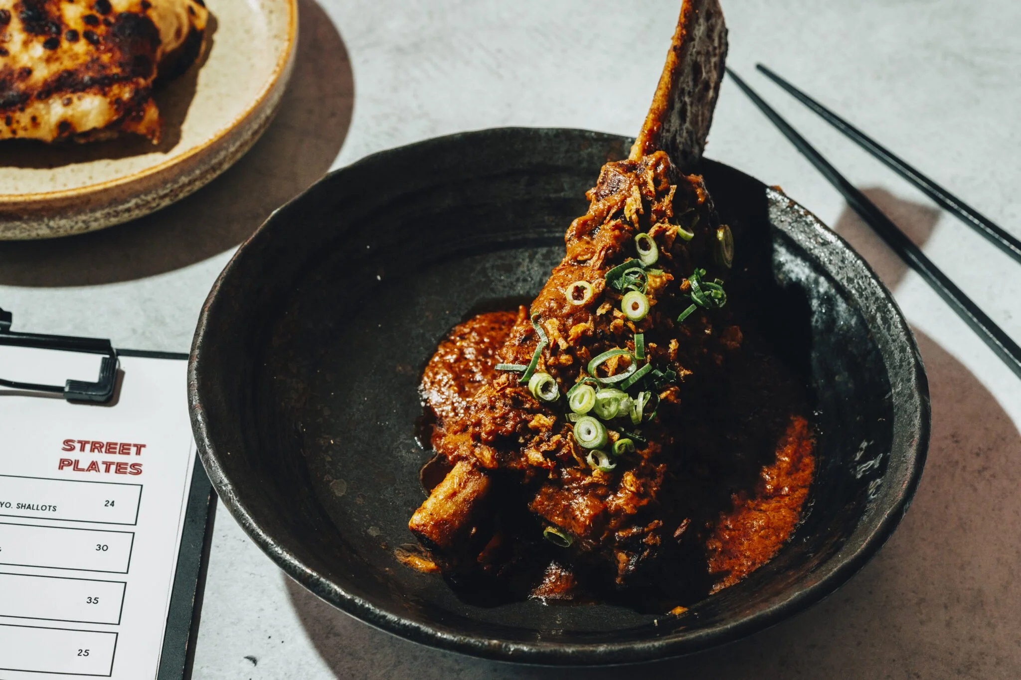 A black bowl containing a braised beef short rib with sauce, garnished with sliced green onions, on a gray table. There are black chopsticks and a plate with grilled chicken in the background.