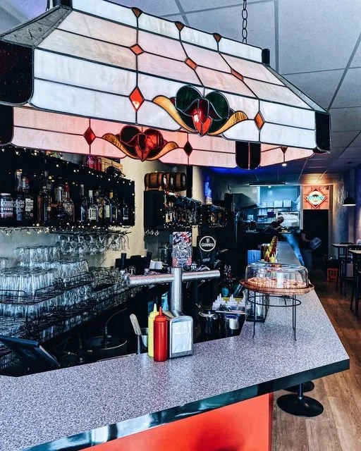 A neon-lit bar with a stained glass light fixture overhead, shelves of liquor bottles, glasses, and bar equipment, and a cozy dining area in the background.