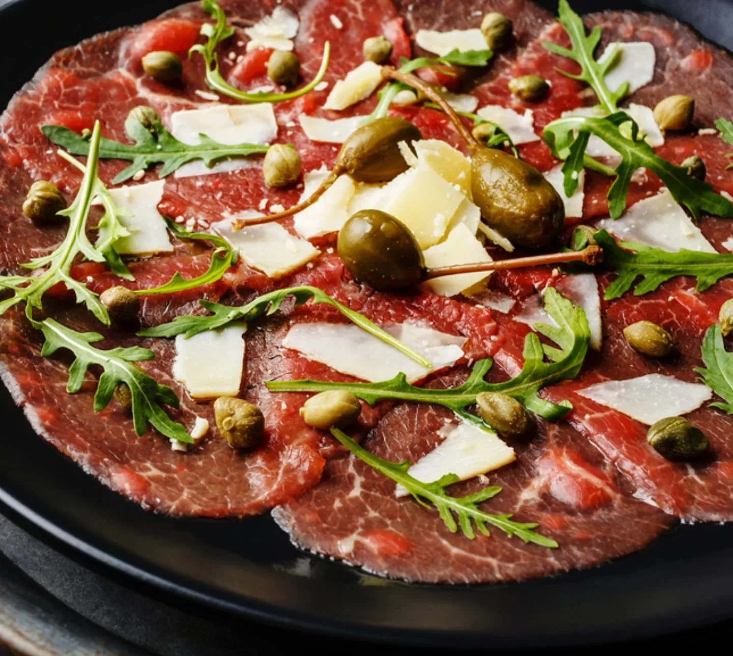 Thinly sliced raw beef topped with arugula, capers, Parmesan cheese shavings, and olive oil on a black plate.