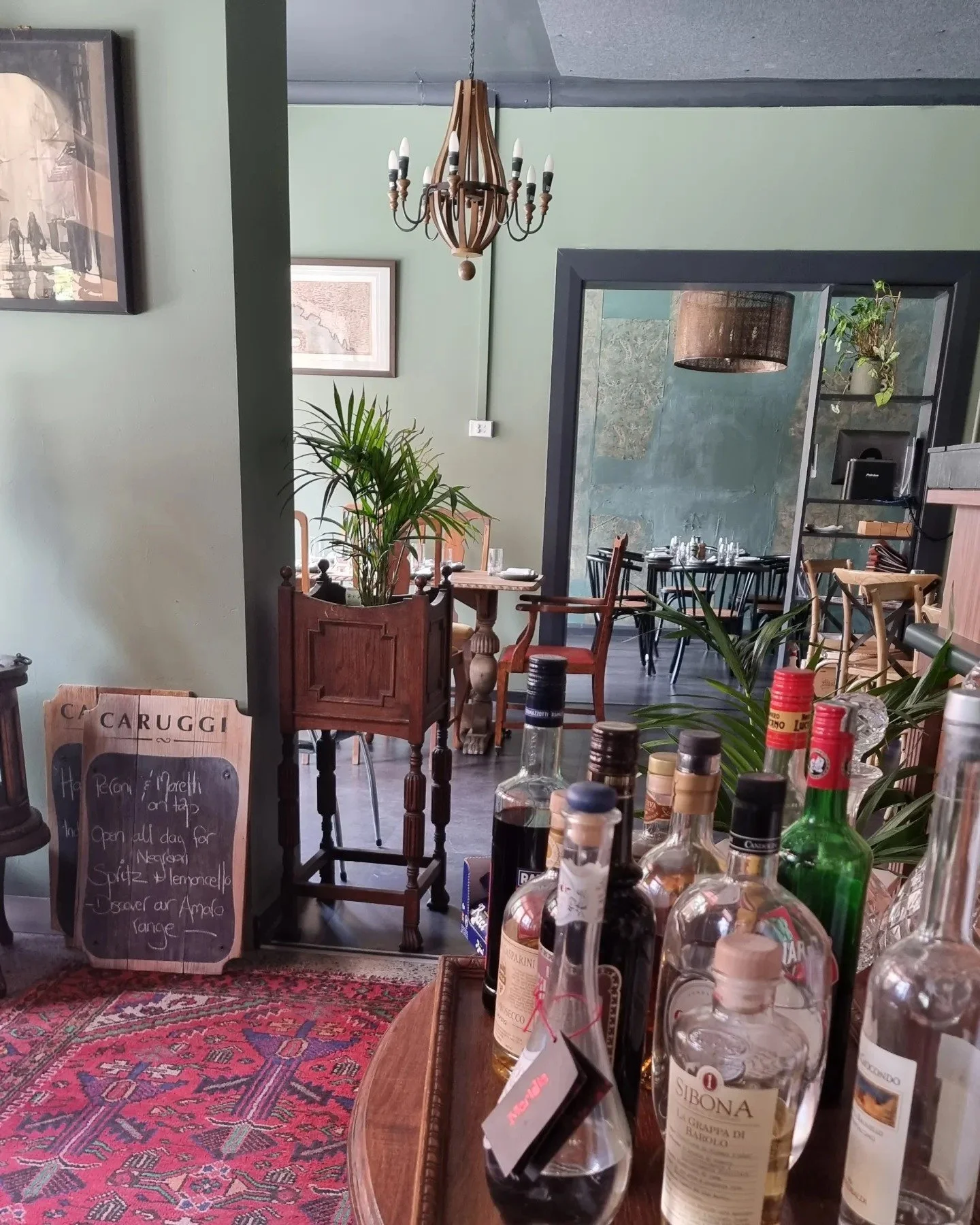 Interior of a cozy restaurant with a display of liquor bottles in the foreground, green walls, wooden furniture, plants, and a chandelier hanging from the ceiling.