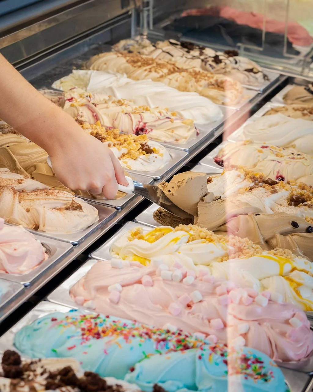 A person's hand scooping strawberry cheesecake ice cream from a display case filled with various flavors of ice cream topped with sprinkles, marshmallows, and candies.