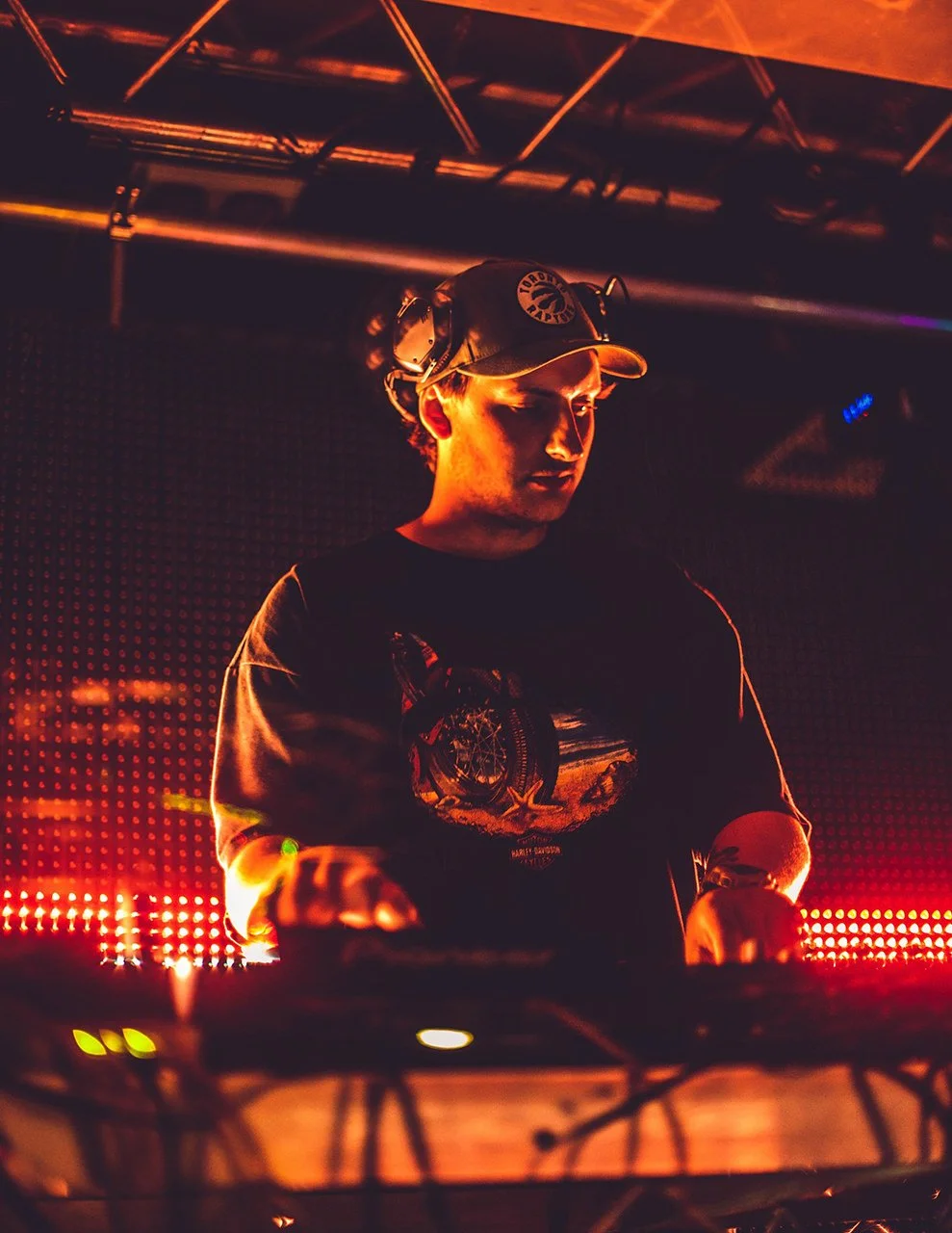 A DJ wearing a cap and headphones performing in a dark club with orange and red lighting.