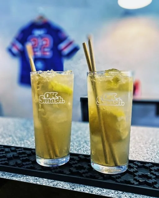 Two tall glasses of yellow-colored drinks with ice and lime wedges, placed on a bar mat, with a blurred background showing a jersey hung on the wall.