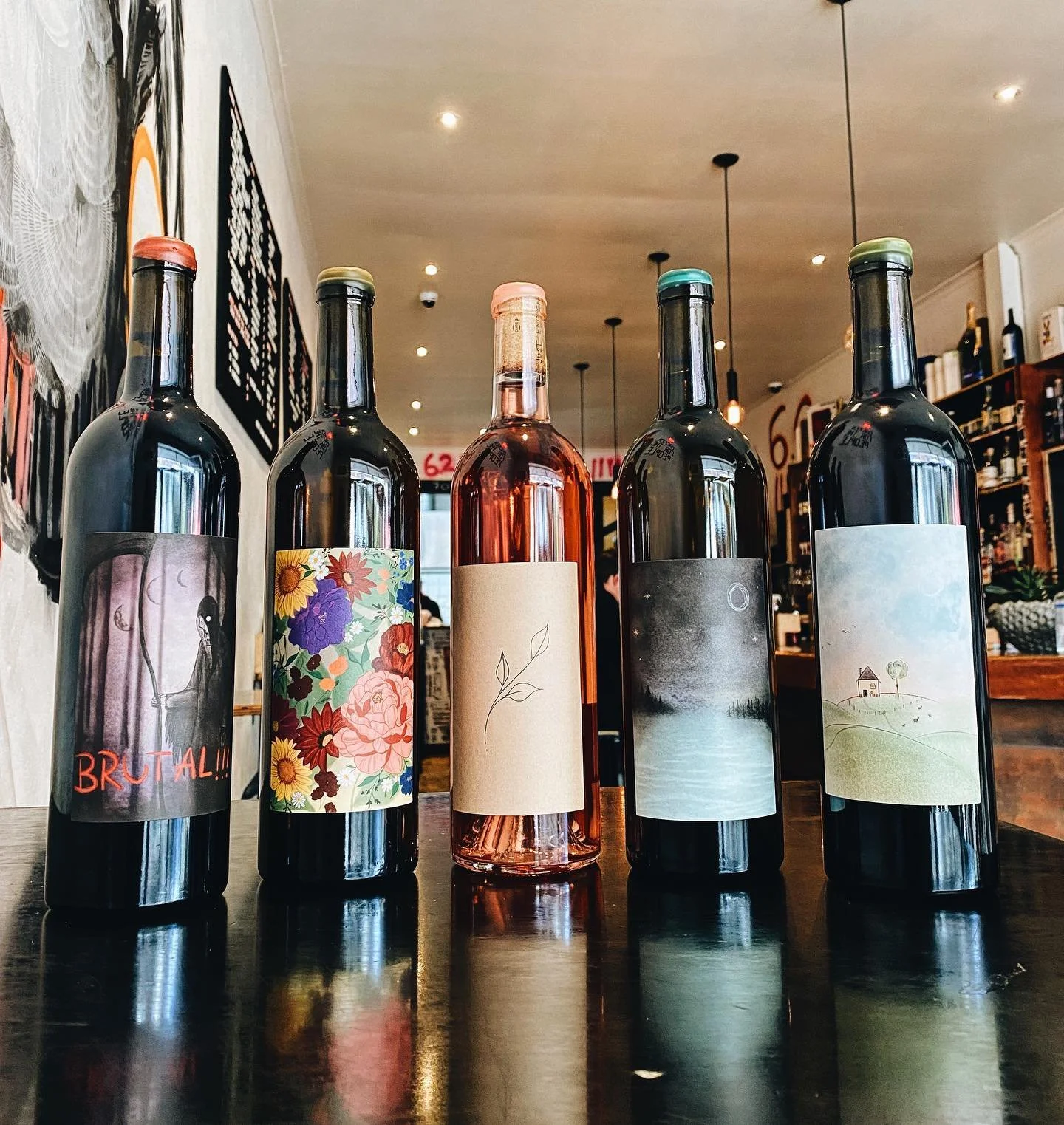Five bottles of wine with different labels on a dark wooden table inside a cozy wine shop or bar. The background shows shelves with more bottles and a few customers.