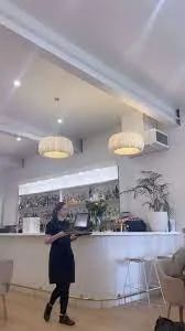 A woman standing at a white bar counter inside a modern, well-lit cafe or restaurant with hanging pendant lights and potted plants, preparing to order or serve.