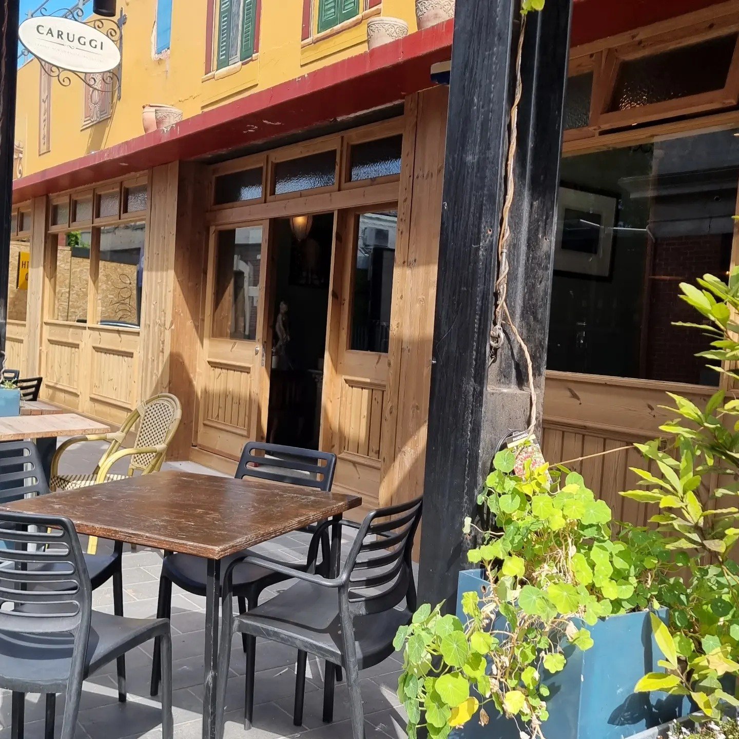 Outdoor seating area with a wooden table and black plastic chairs, next to a building with wooden doors and large windows, and a bright yellow painted wall with an embedded colorful mural. A blue planter with green plants is in the foreground.