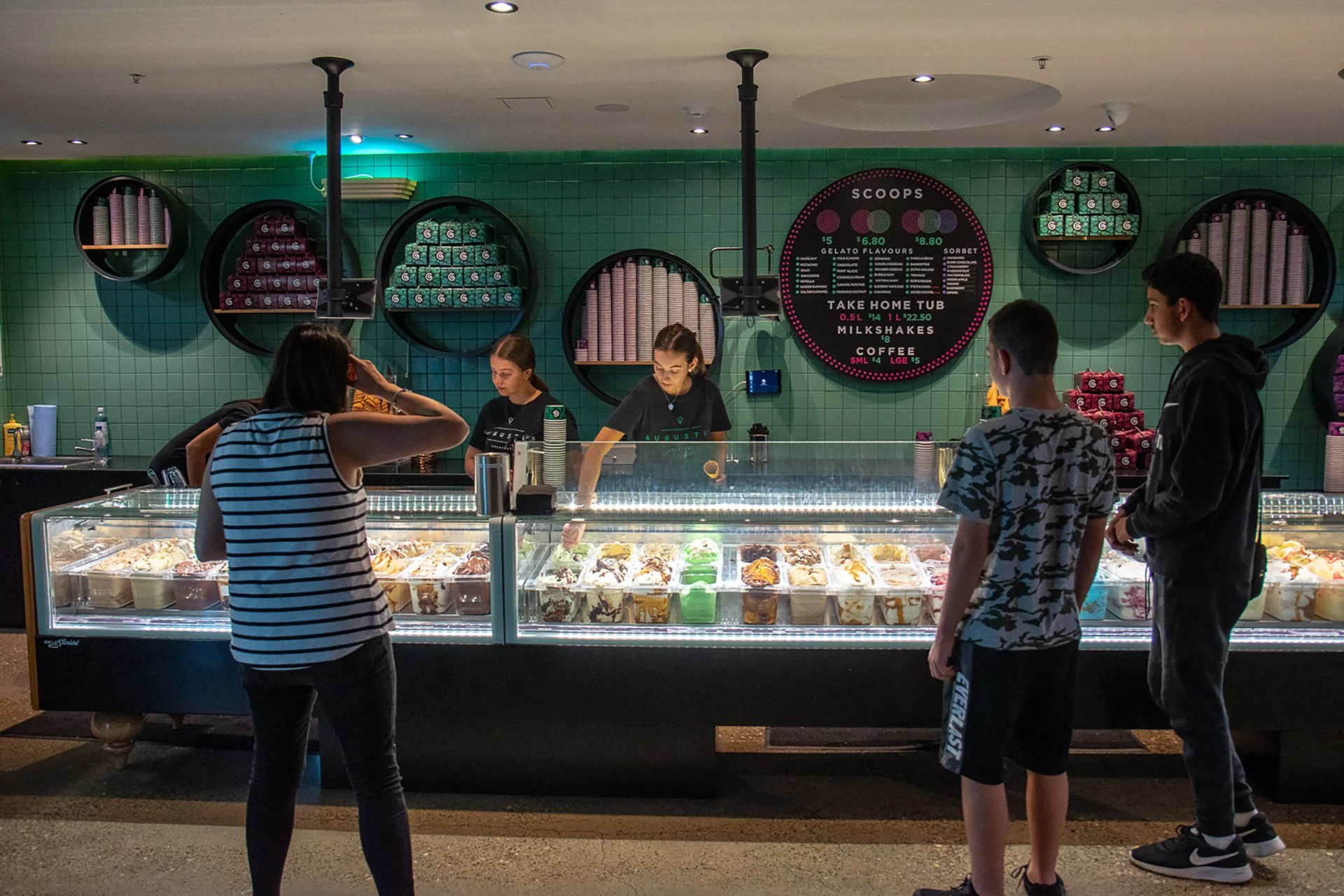 People standing in front of an ice cream shop counter with three employees serving ice cream. The shop has a green-tiled wall with circular shelves holding cups and flavor containers, and a menu board.