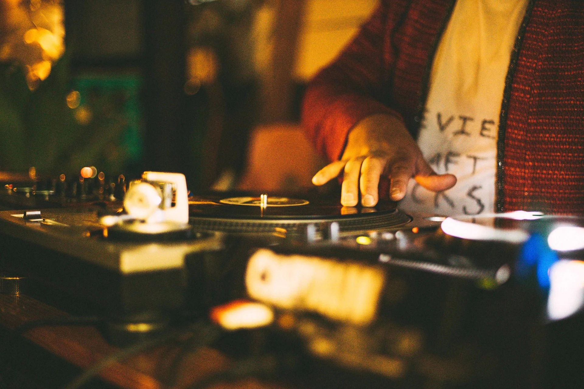 A DJ's hand on a turntable in a dimly lit environment with warm, ambient lighting.