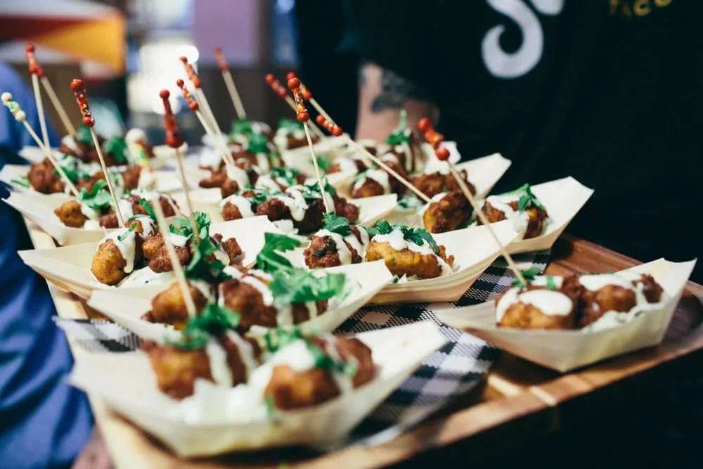 Several small paper boats filled with fried food bites topped with white sauce, green herbs, and colorful toothpick toothpicks, arranged on a wooden tray.
