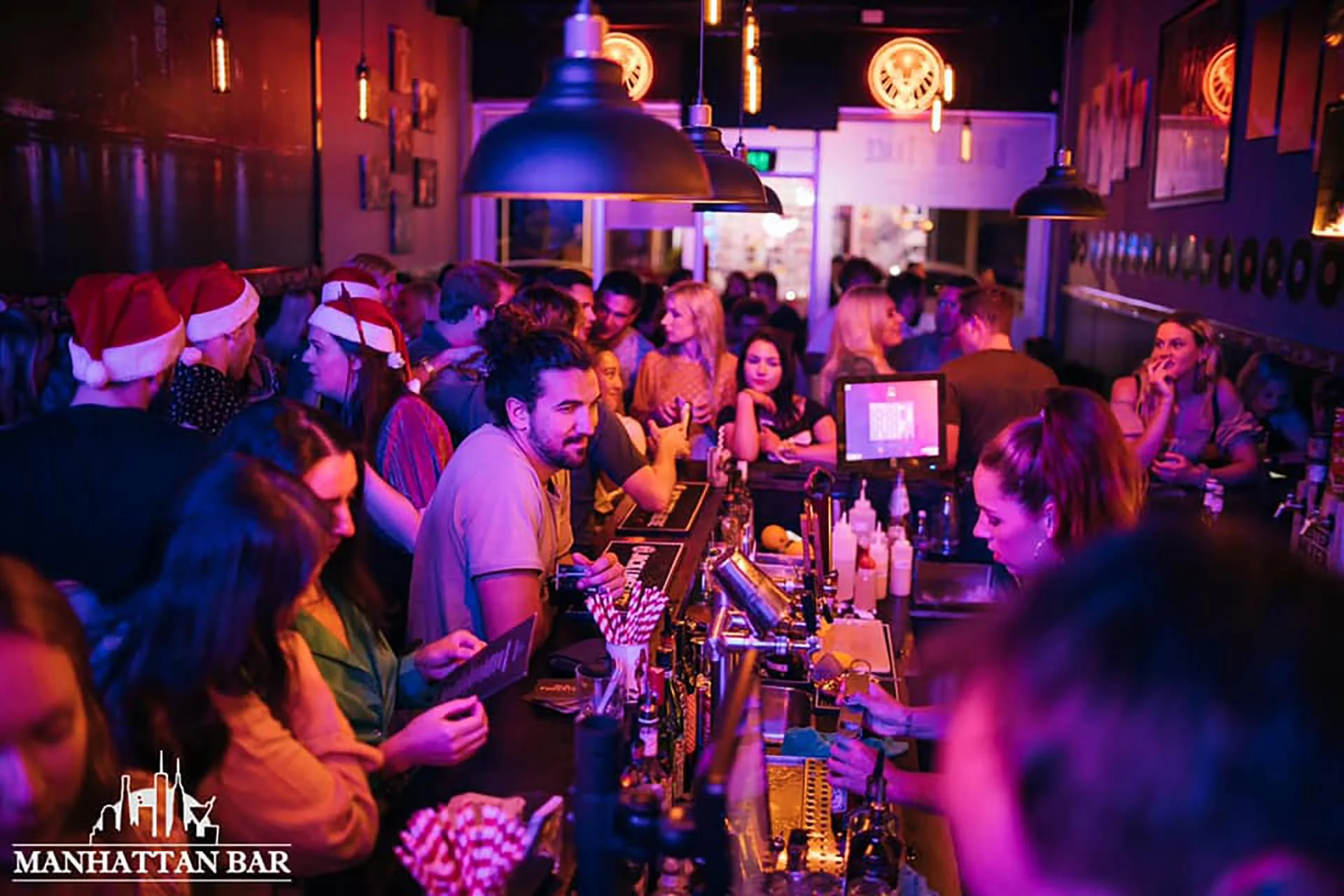 A lively bar scene with many people, some wearing Santa hats, socializing and enjoying drinks. The bar has colorful lighting and decorations, with staff serving at the counter. The logo in the bottom left corner reads 'Manhattan Bar'.