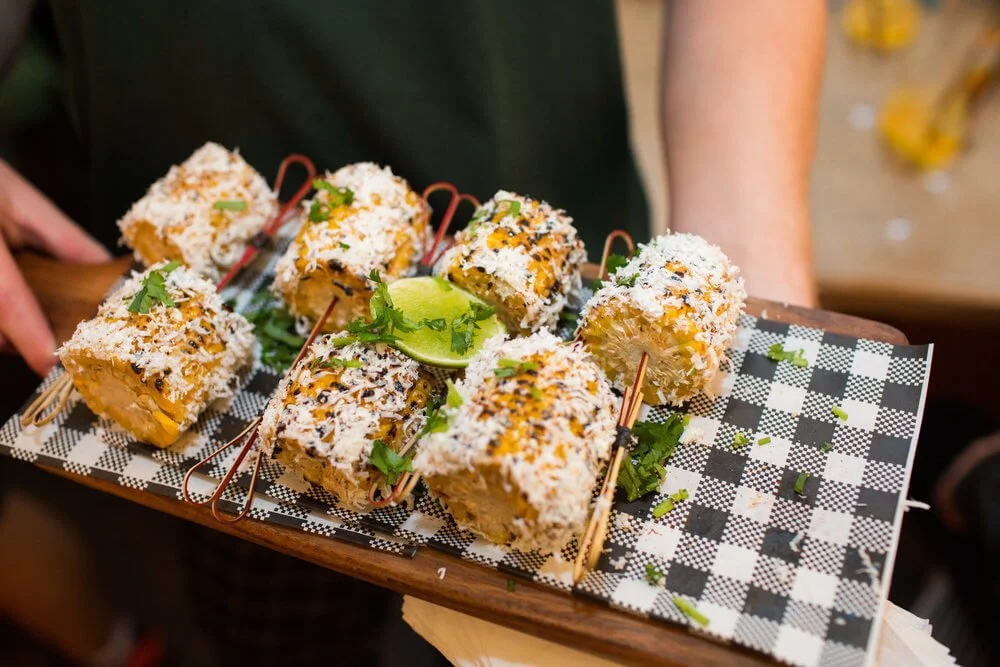 Tray of seven coconut-covered fried rolls garnished with cilantro and lime wedge on checkered paper.