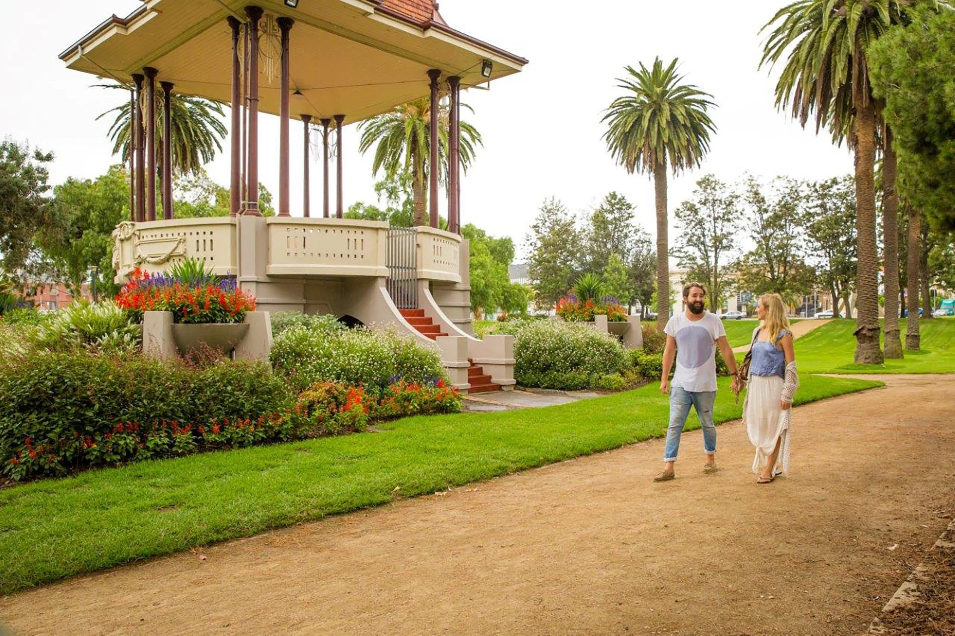A man and woman walking hand in hand on a dirt path in a park with green grass and tall palm trees, with a decorative bandstand in the background surrounded by colorful flowers.