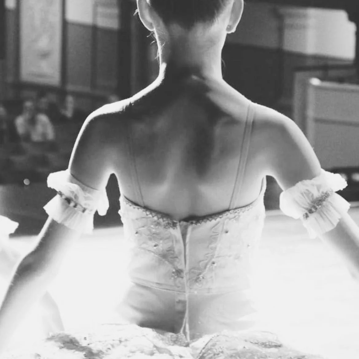 Black and white photo of a ballet dancer from behind, wearing a corset-style costume with ruffled sleeves, in a dance studio or classroom setting.
