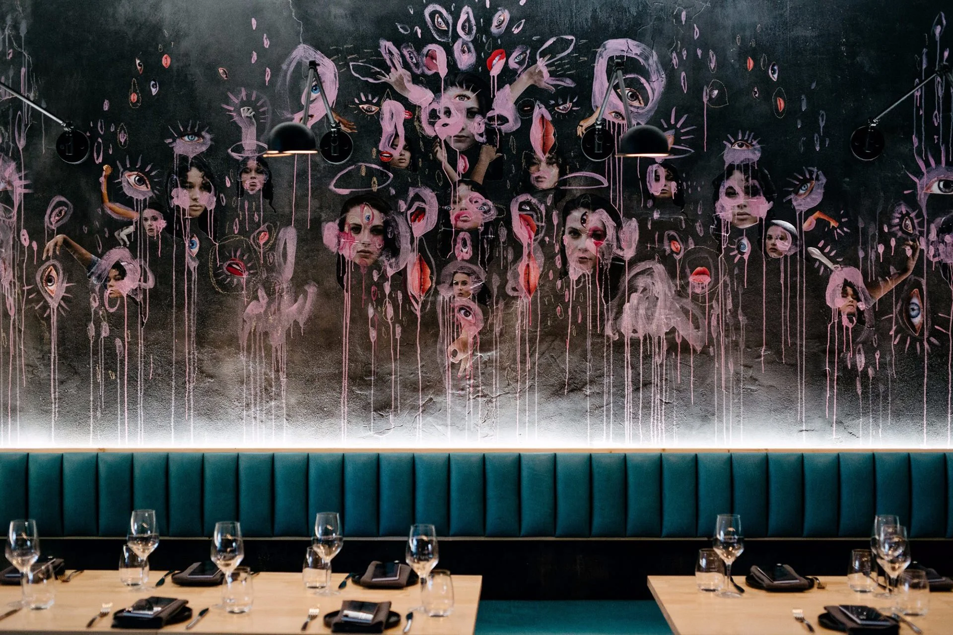 A mural featuring multiple human faces and eyes painted on a dark wall above a teal banquette in a restaurant, with tables set with wine glasses, utensils, and black napkins.