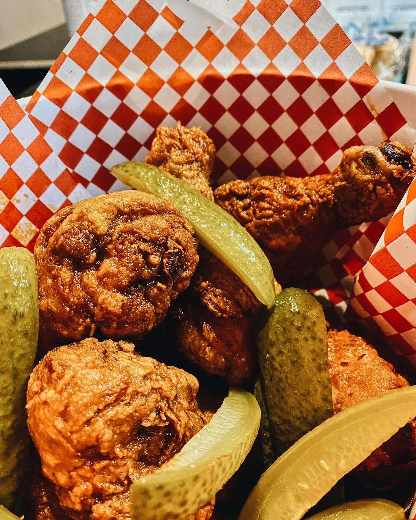 Close-up of fried chicken, meatballs, pickles, and lemon wedges in a red and white checkered paper basket.