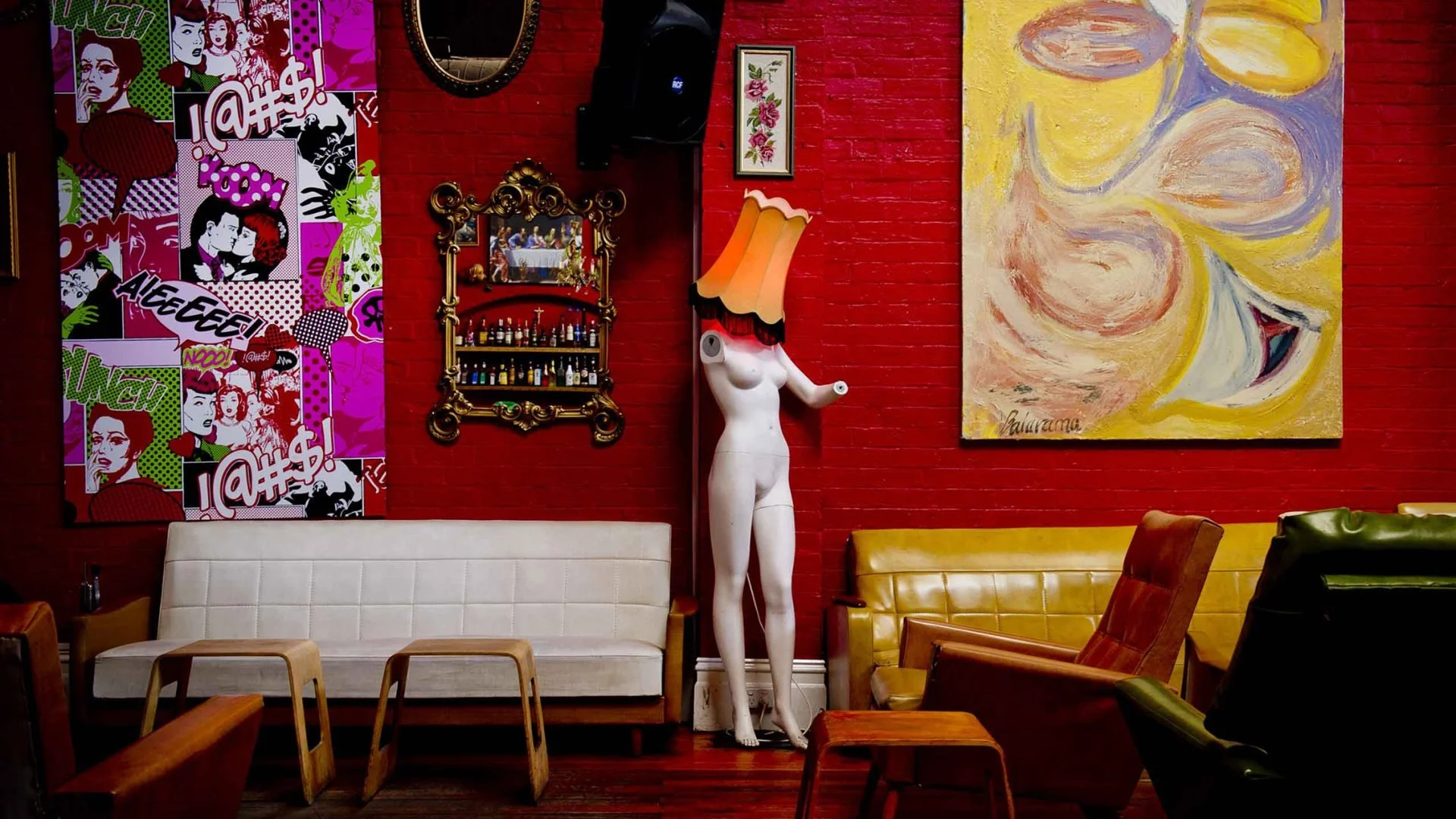 Interior of a colorful cafe or lounge with a variety of eclectic art and furniture. The wall features a bright red brick background decorated with pop art, a framed picture, a decorative mirror, a sculpture of a headless woman with a lampshade as her