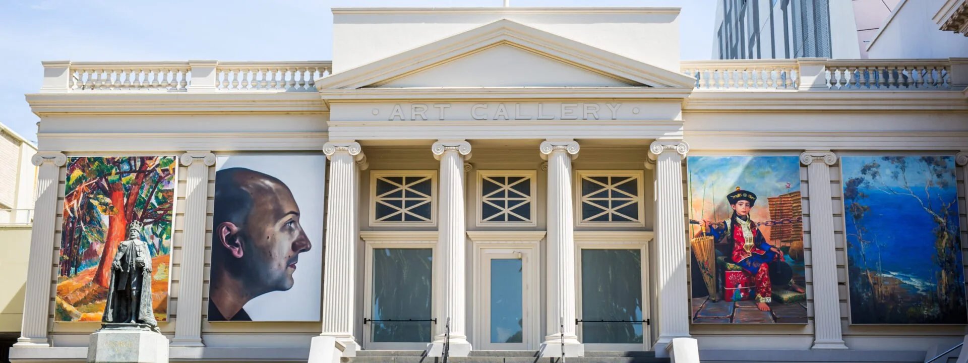 Exterior of an art gallery building with large paintings on display, columns, and a statue in front.