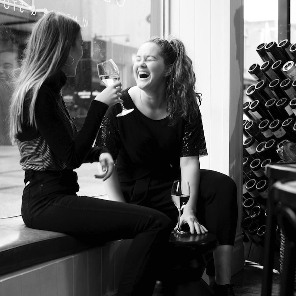 Two women sitting on a bench inside a wine store, laughing and holding glasses of wine, with wine bottles on a rack behind them.
