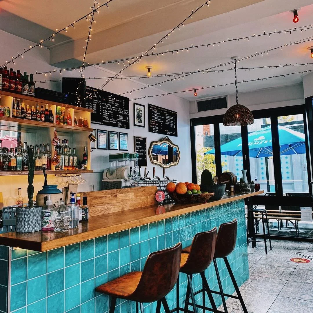 Interior of a cozy cafe with a wooden bar counter, turquoise tiled front, bar stools, and hanging string lights on the ceiling. Inside, there are liquor bottles on shelves, a coffee machine, and a bowl of fruit on the counter. Large windows with a vi