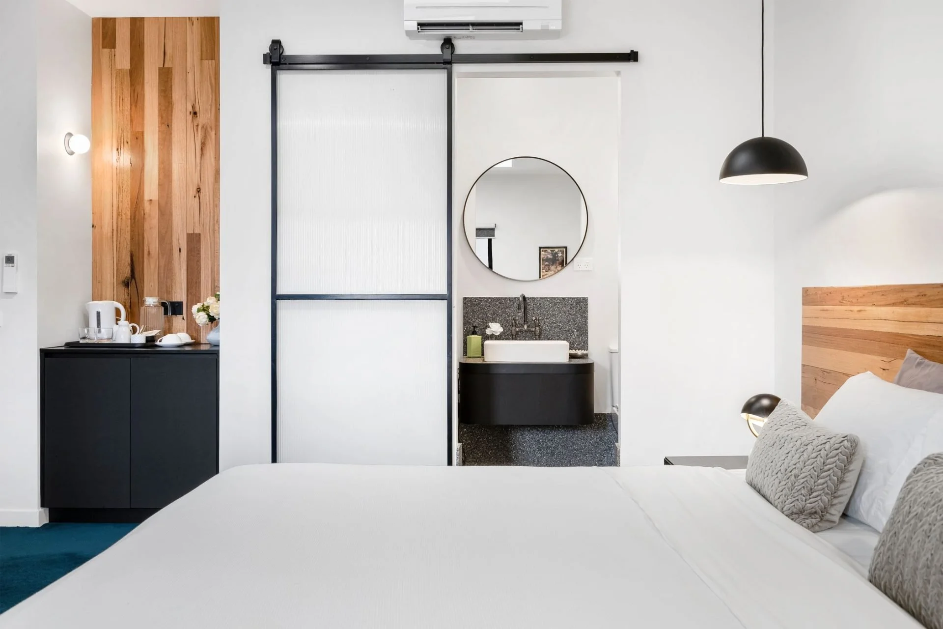A bedroom with a white bed, gray pillows, a black pendant light, and a black butler sink with a round mirror, a soap dispenser, and a flower. A sliding door separates the bedroom from a small kitchenette with a black cabinet, a white electric kettle,