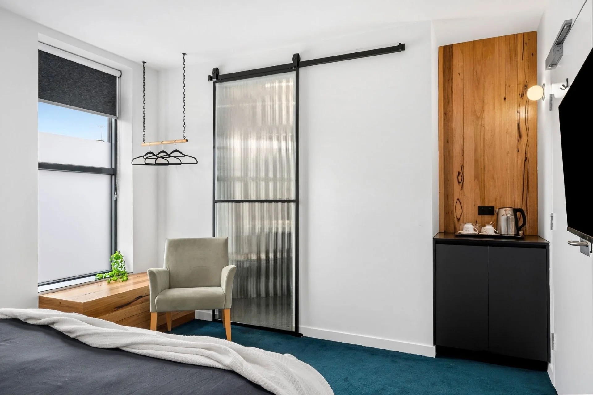 Modern bedroom with large window, beige armchair, wooden accents, black cabinet with tea set and electric kettle, frosted glass sliding door, and flat-screen TV.