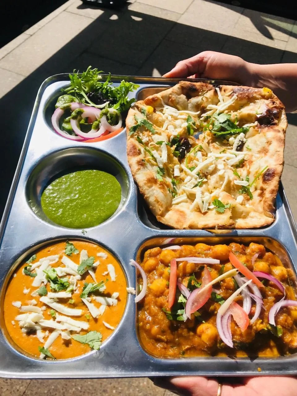 A stainless steel meal tray containing a slice of pizza, two bowls of curry, a small container of green chutney, and a side salad with onion rings and cilantro.