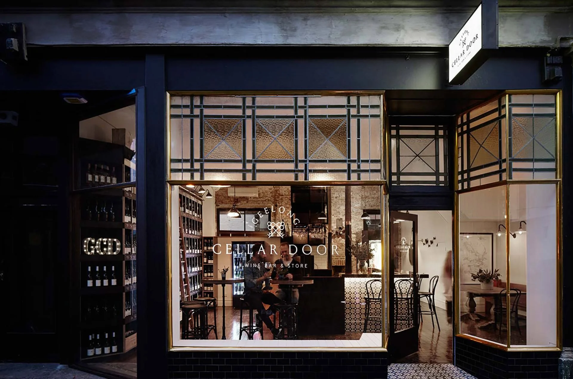 Exterior view of Cellar Door wine bar and store with large glass windows, showing inside seating and wine shelves, and a sign reading 'Cellar Door' above the entrance.
