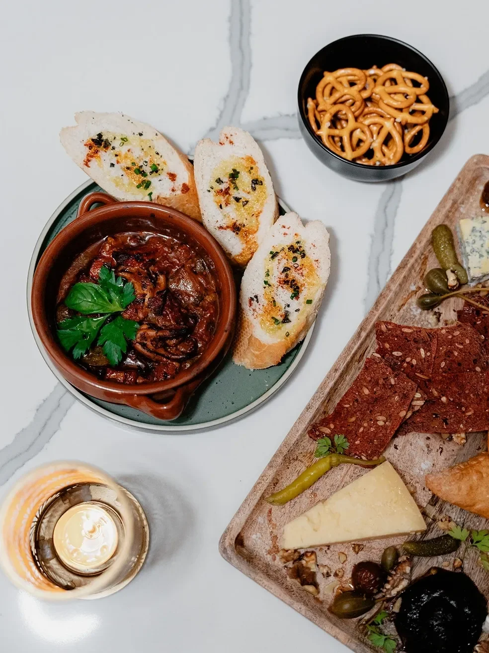 A serving of Italian antipasto includes toasted baguette slices with garlic and herb spread, a small bowl of pretzels, a ceramic dish of braised meat with herbs, and a wooden platter of cheese, crackers, and cured meats with garnishes, all on a white
