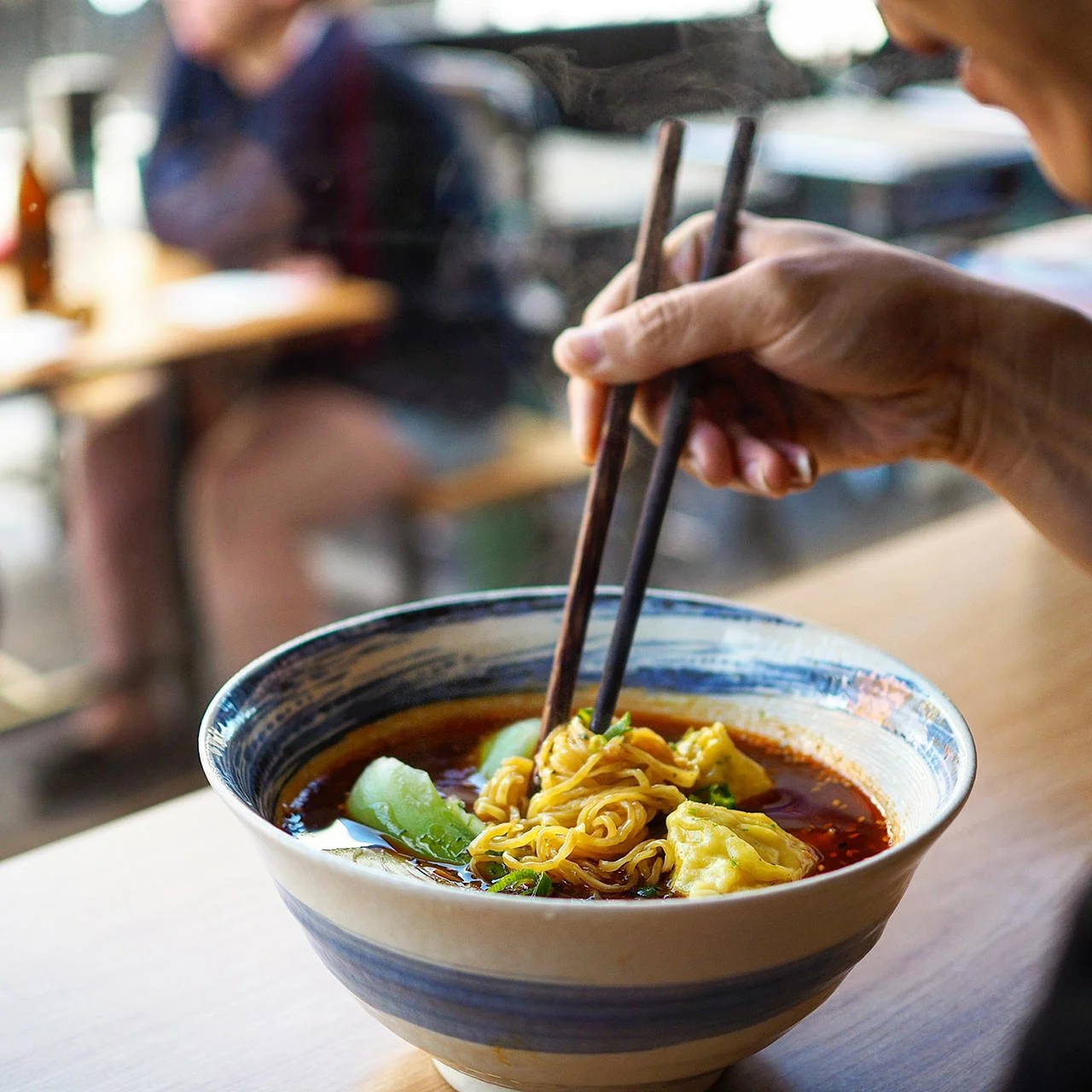 Person using chopsticks to eat noodles and dumplings from a bowl of spicy soup in a restaurant.