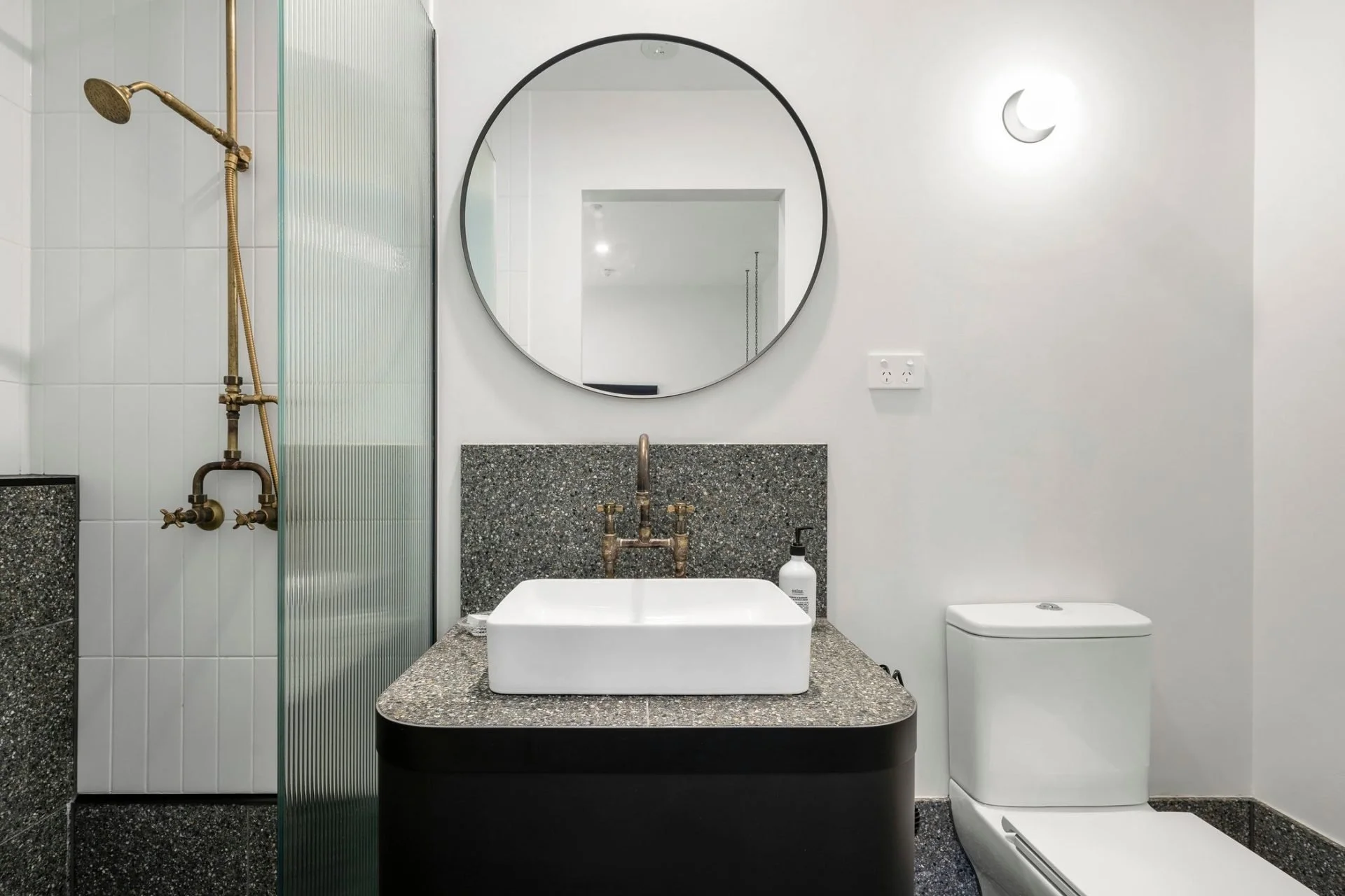 Modern bathroom with a white square vessel sink on a speckled black countertop, a round mirror, a crescent moon-shaped wall light, a toilet, and a shower with a brass showerhead and fixtures, all set against white and speckled black walls.