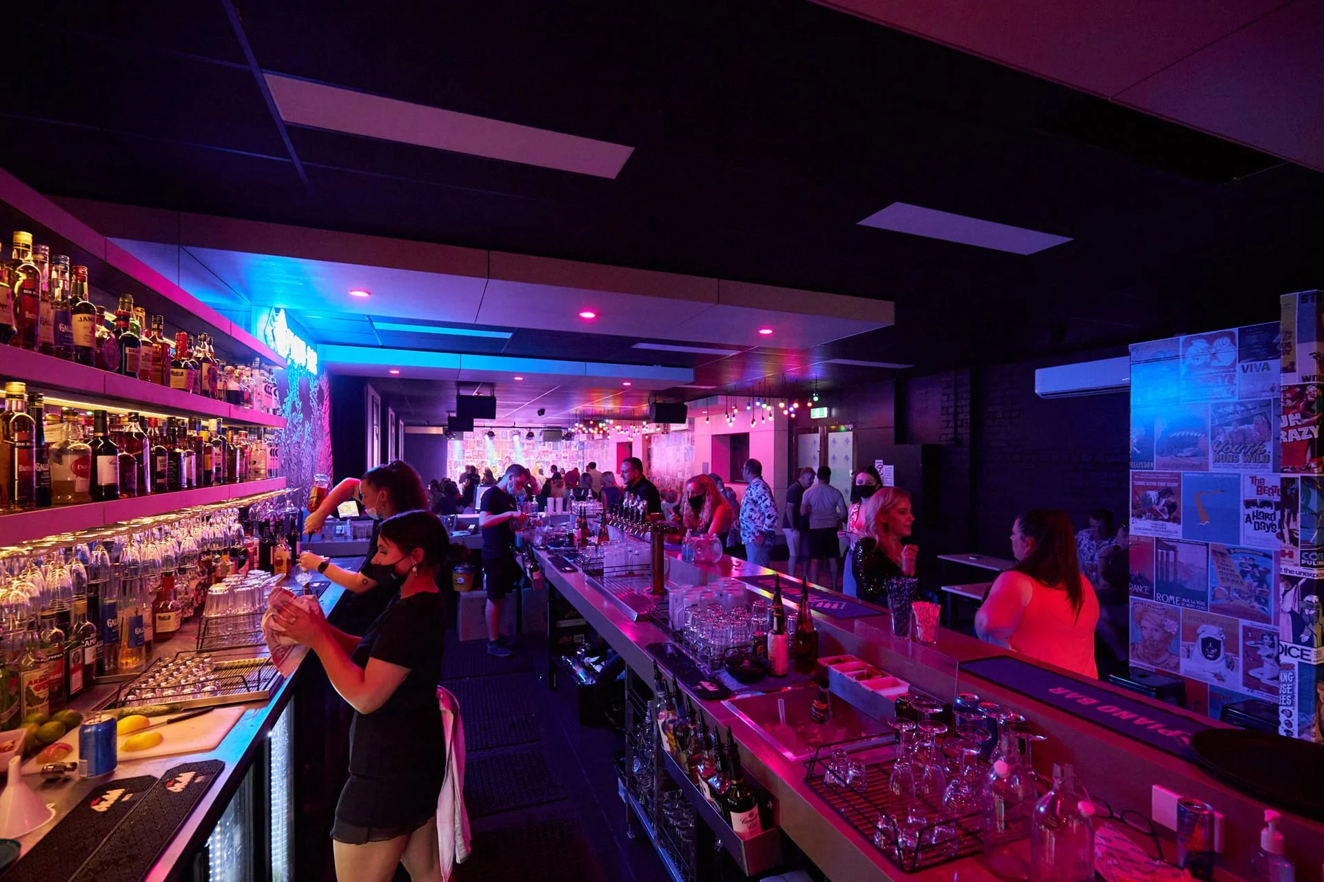 A busy nightclub bar with bartenders preparing drinks and patrons socializing under colorful lighting.