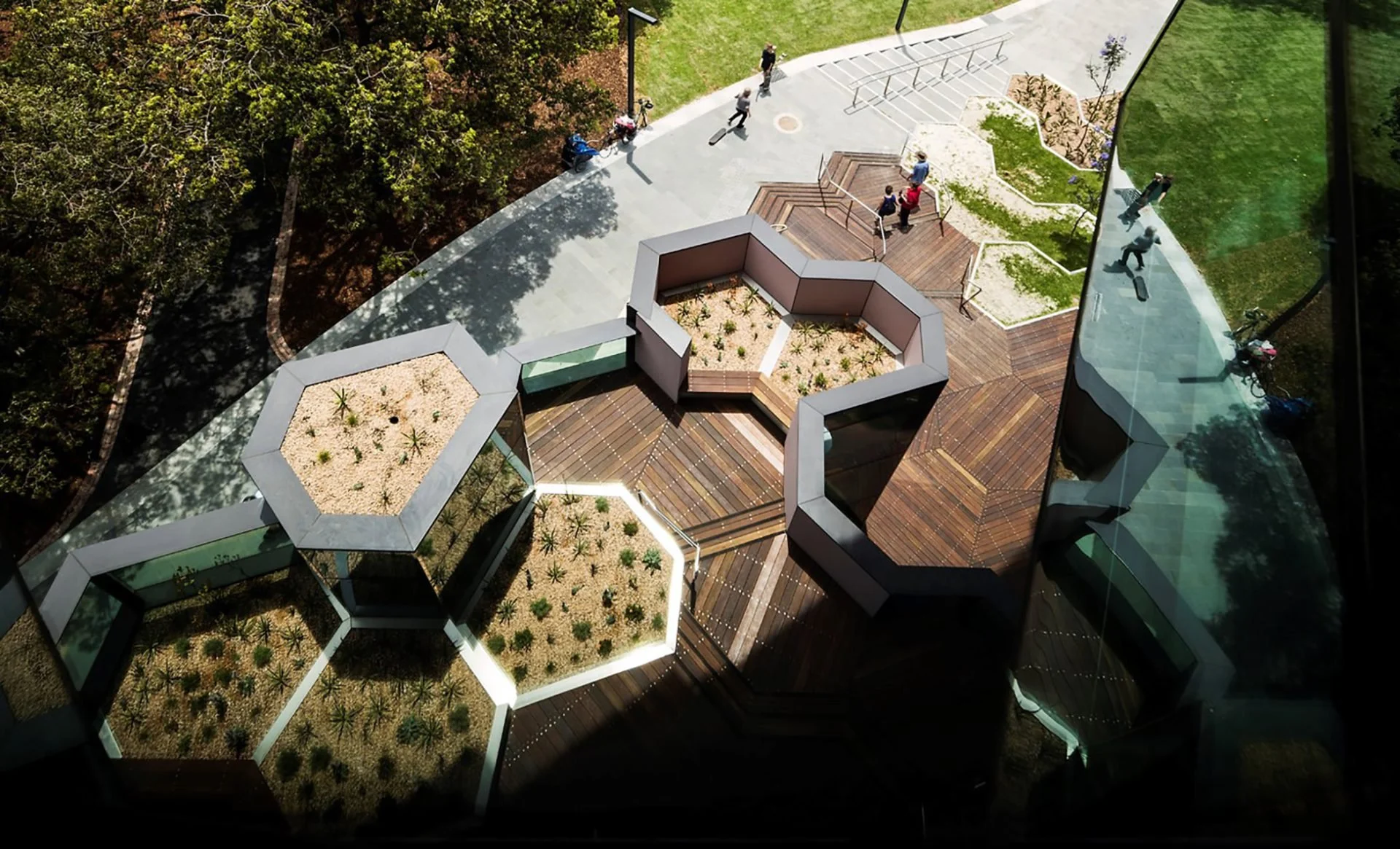 An aerial view of a modern urban outdoor space featuring hexagonal planter beds with plants, a wooden deck with stairs, and a glass reflective building wall. Several people are walking on the paved path beside the garden area.