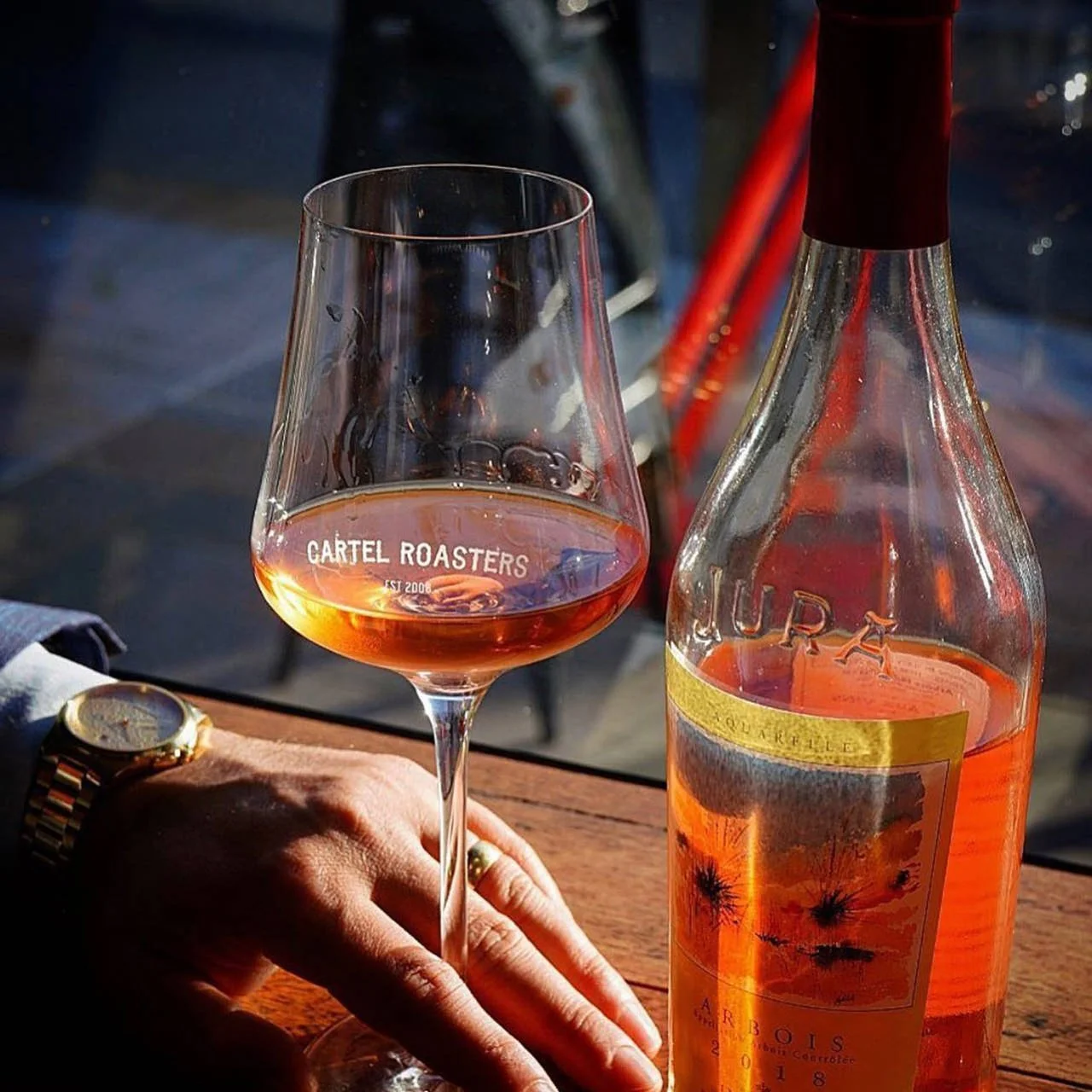 A glass of rosé wine and a bottle on a wooden table, with a person's hand partially visible, wearing a watch and a ring, in a setting with sunlight and a blurry background.