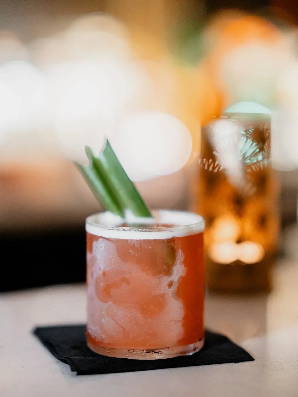 A close-up of a cocktail in a short glass with a frosted exterior, garnished with green drink stirrers, placed on a black napkin.