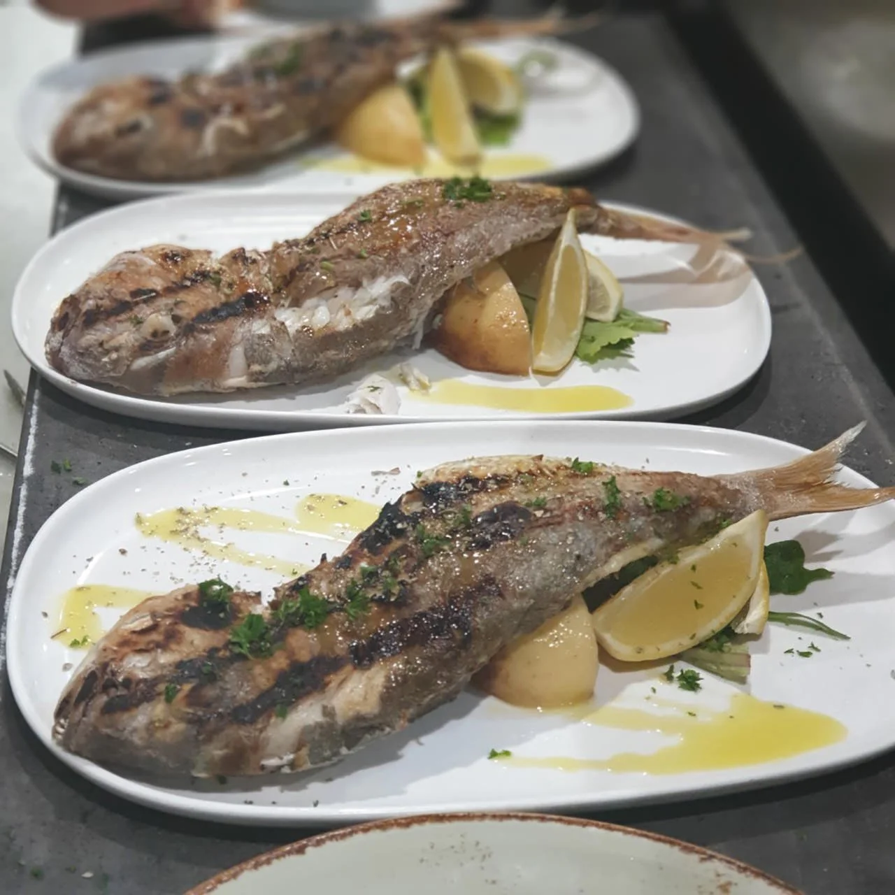 Three plated grilled fish with lemon wedges, herbs, and drizzled olive oil on white plates.