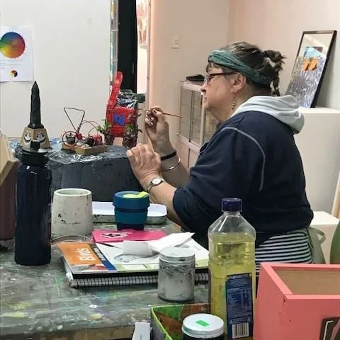 Woman sitting at a cluttered table, painting a small object, with art supplies, books, and cups on the table, and framed artwork leaning against the wall in the background.