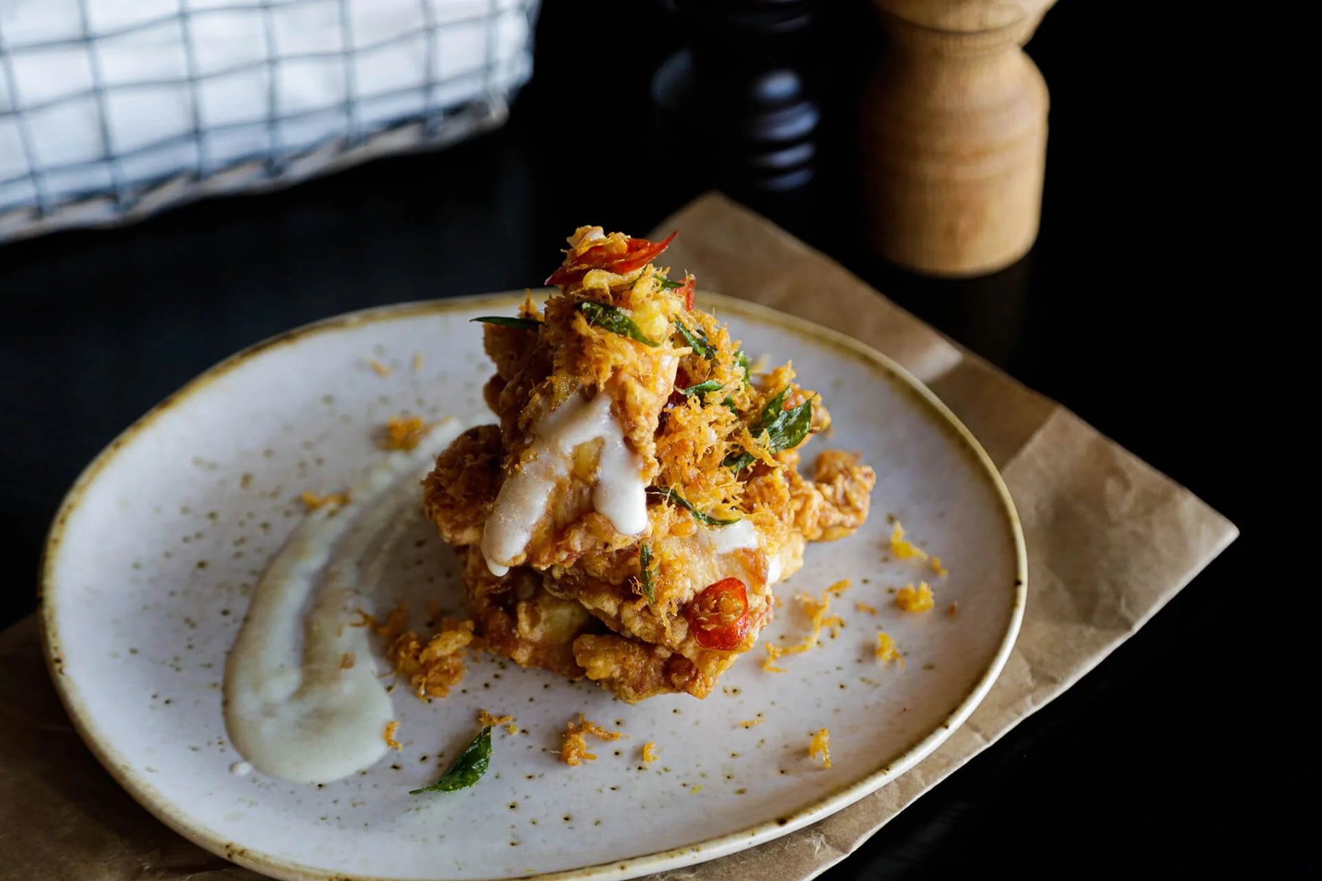 Fried chicken topped with herbs and chili, served with a drizzle of white sauce on a white plate with gold rim on brown parchment paper.