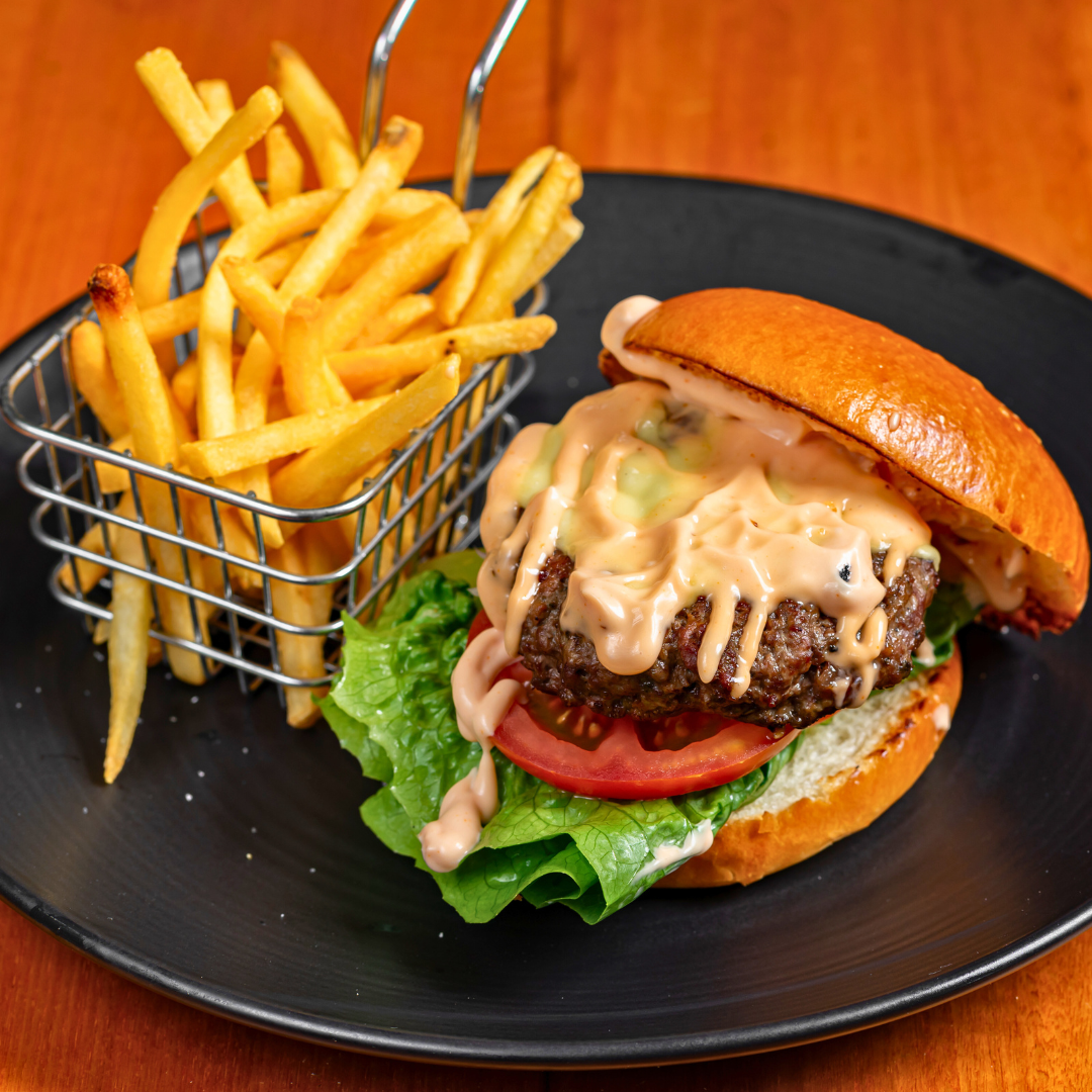 Plate with French fries in a small metal basket and a cheeseburger with lettuce, tomato, beef patty, cheese, sauce, and sandwich bun on a black plate.