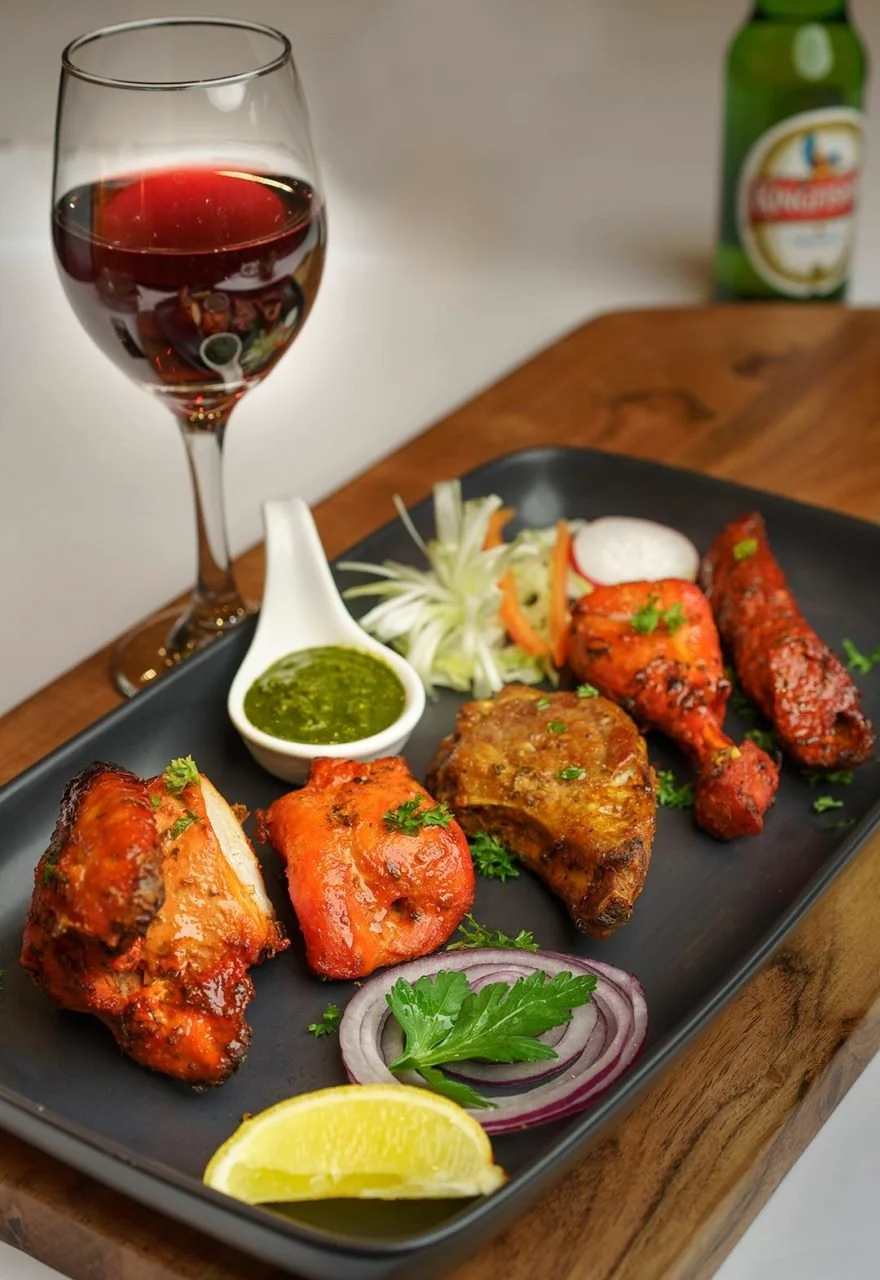 A black rectangular plate with four pieces of spicy grilled chicken drumsticks garnished with cilantro, served with sliced onions, lemon wedge, and a small bowl of green chutney. A glass of red wine and a bottle of beer are in the background on a woo