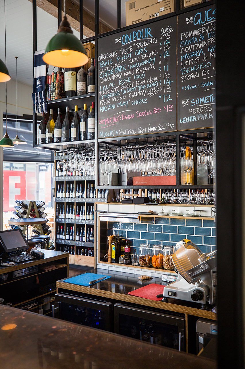 A bar counter with wine bottles, hanging glasses, a chalkboard menu with wine selections, and a meat slicing machine.