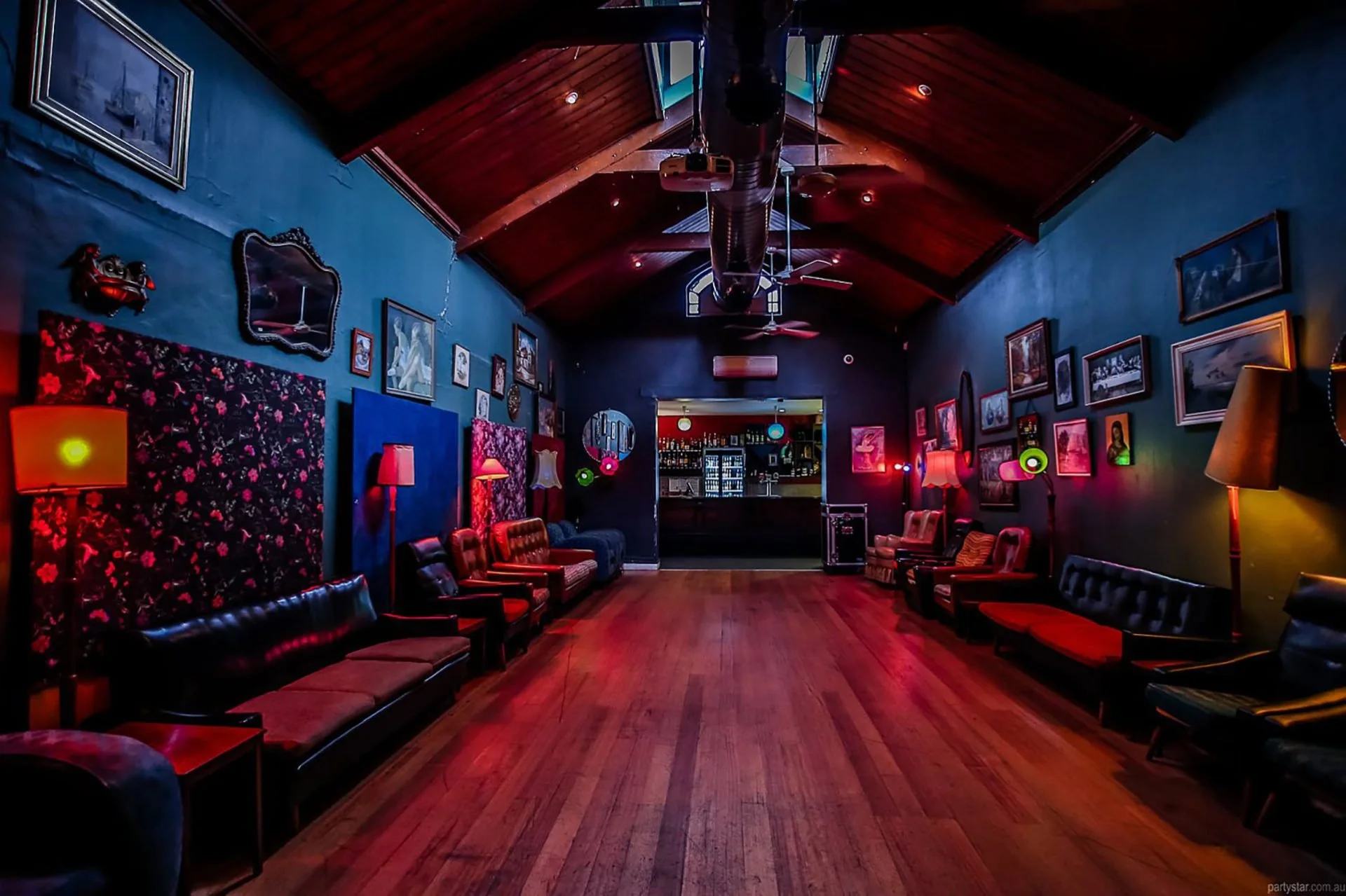 A cozy, dimly lit lounge with vintage decor, framed pictures on dark walls, and a wooden ceiling. Multiple couches and chairs are arranged along the walls, with lamps providing a warm glow. A bar area is visible in the background.