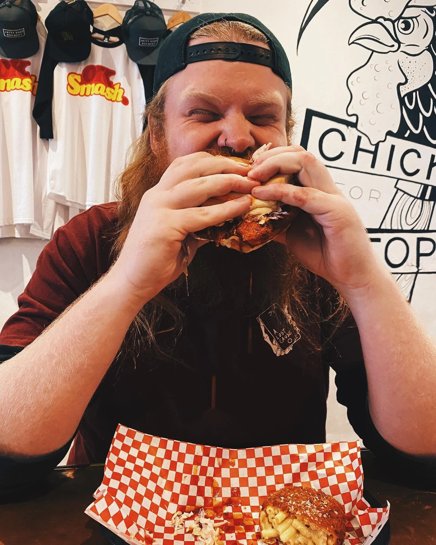 Man with long hair and beard, wearing a backward cap, enjoying a large sandwich in a restaurant with chicken-themed decor.
