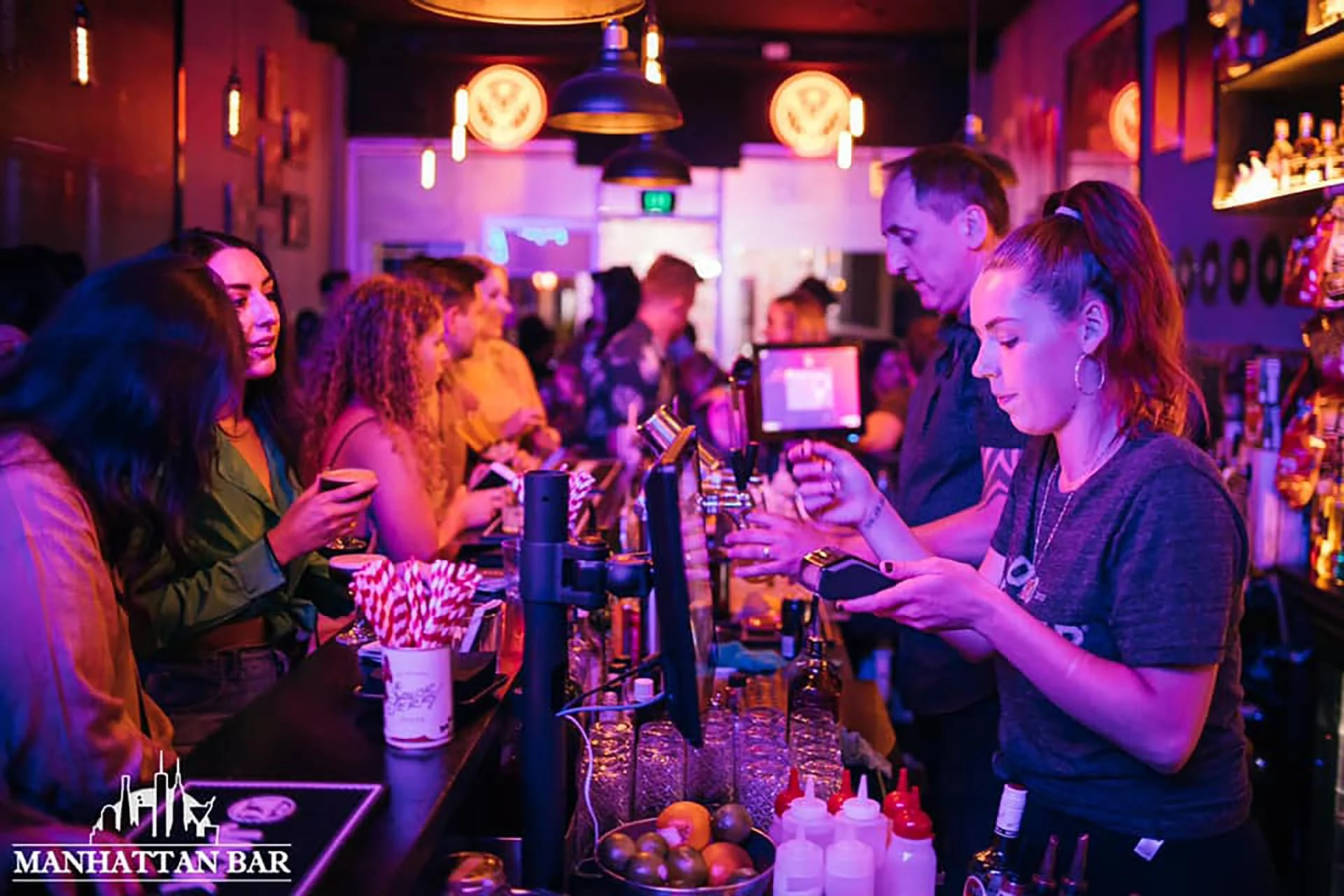 Group of people at a bar with neon lights, some are ordering drinks, bartender preparing drinks, colorful decor, and a lively atmosphere