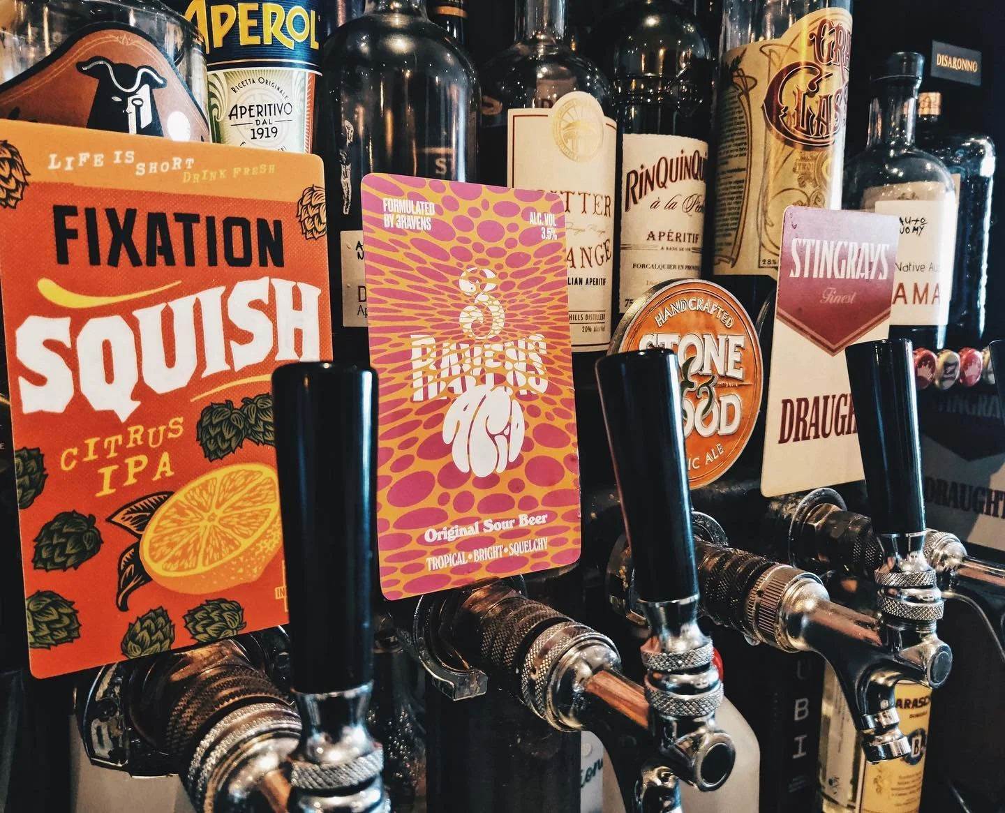 A row of beer taps with colorful labels for various craft beers, including Fixation Squish Citrus IPA, a pink label with a turtle, a Stone & Wood label, and other beer brands behind the taps.