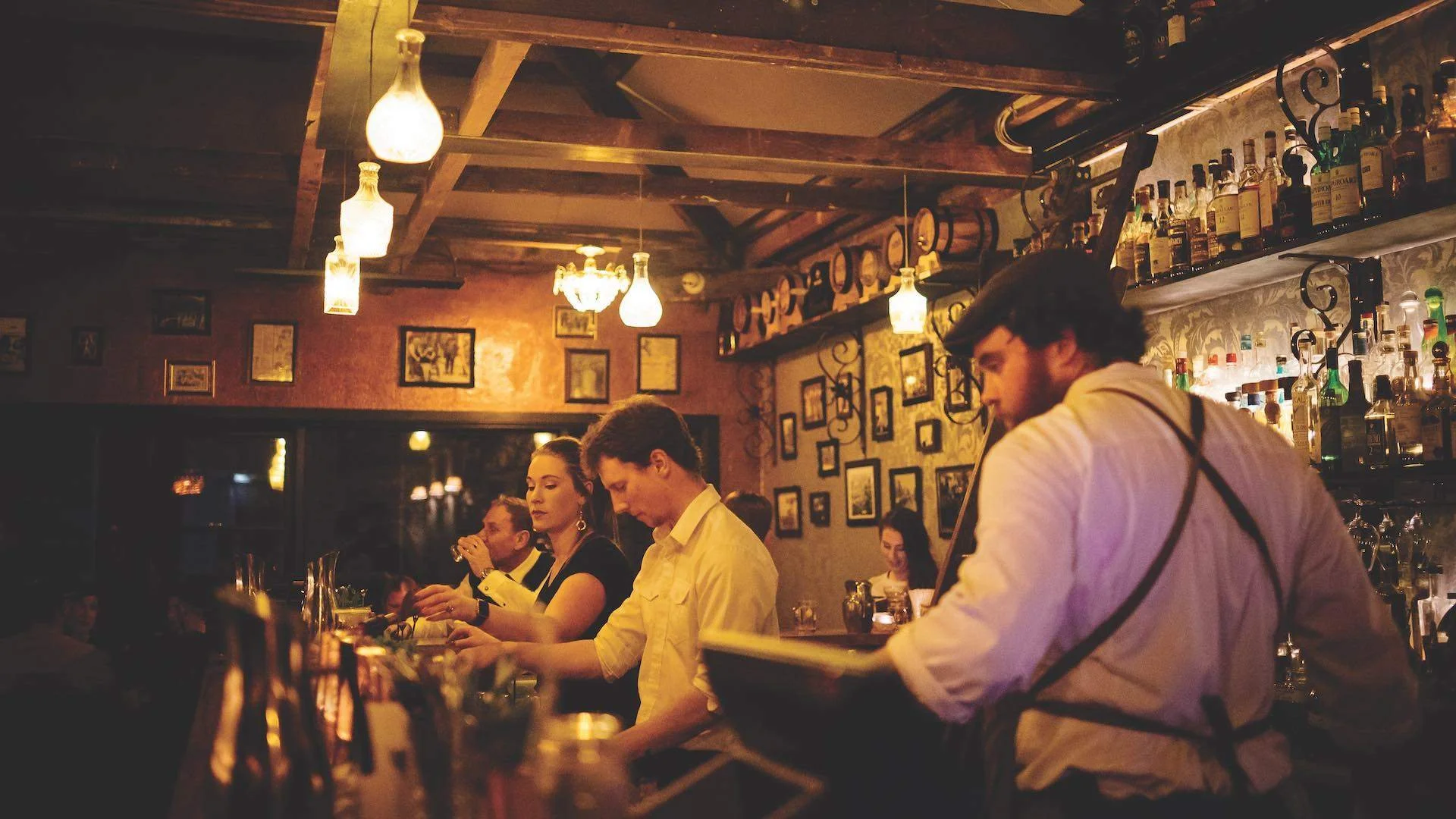 Bar scene in a cozy, dimly-lit restaurant with staff and customers. Three people are sitting at the bar, and a bartender is working behind it. The wall has framed pictures and bottles of alcohol on shelves.