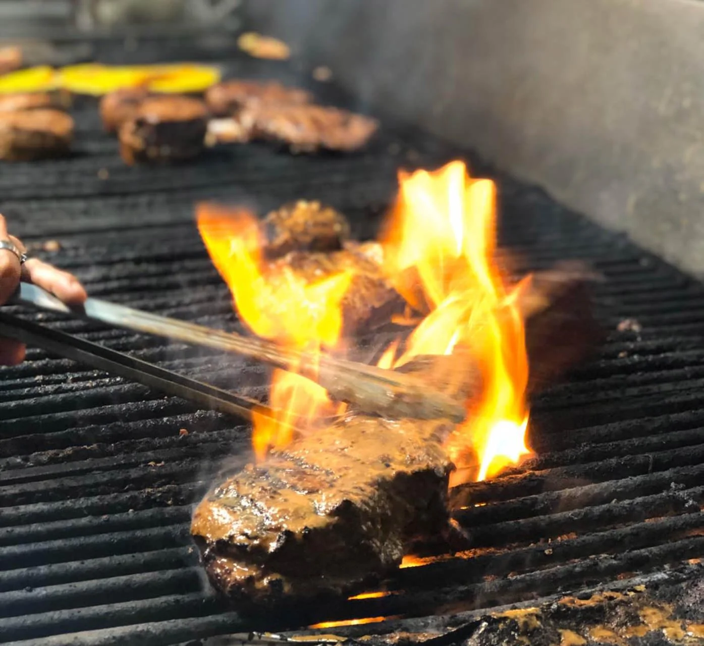 Close-up of a burger patty on a grill with flames, and someone holding tongs near the burger.