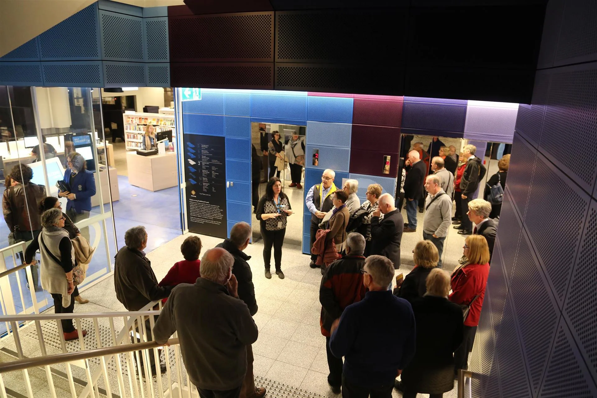 People waiting in line and standing in a lobby with modern blue and purple wall panels, while a woman gives a presentation or speech.