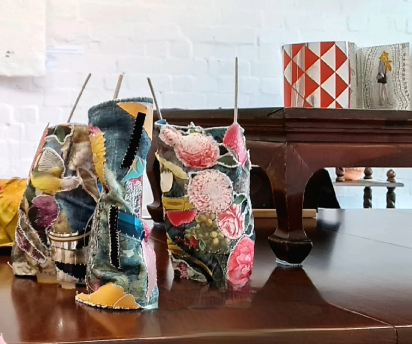 Three fabric vases with floral patterns and a saw inside, placed on a polished wooden table, with decorative boxes and a white brick wall in the background.